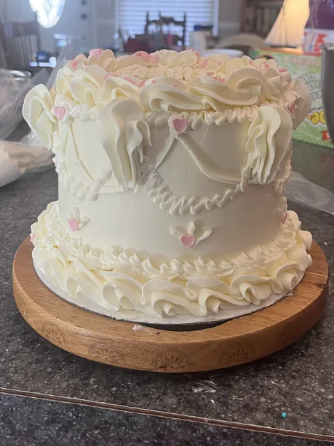 Vintage-style white cake with ruffled piping, piped swags, and small pink hearts on a wooden stand.